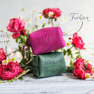 May include: Two small leather pouches with personalized names, "Sadie" and "Marley". The pouch labeled "Sadie" is fuchsia and the pouch labeled "Marley" is forest green. The pouches are surrounded by pink and white flowers.