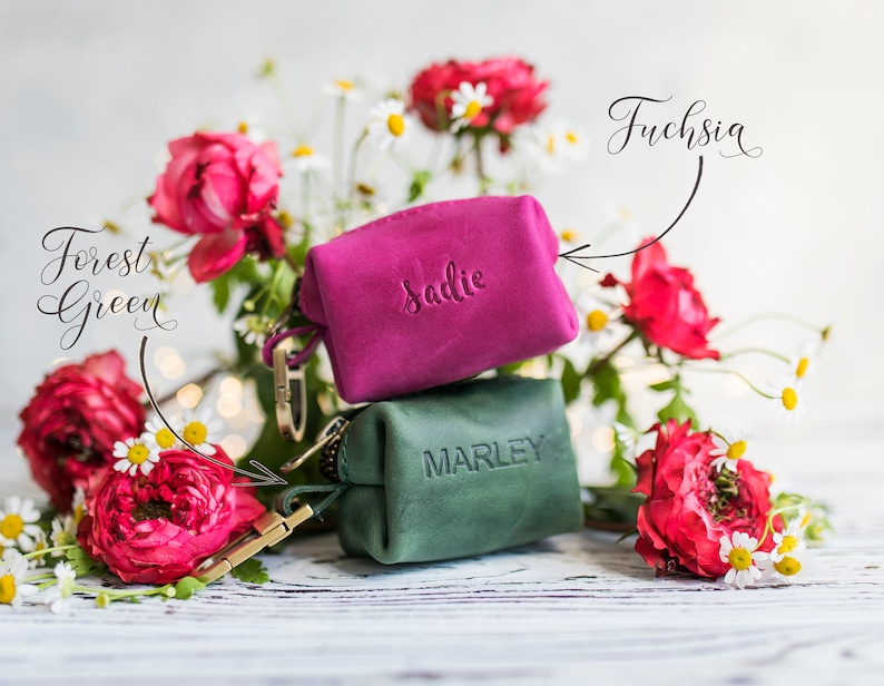 May include: Two small leather pouches with personalized names, "Sadie" and "Marley". The pouch labeled "Sadie" is fuchsia and the pouch labeled "Marley" is forest green. The pouches are surrounded by pink and white flowers.