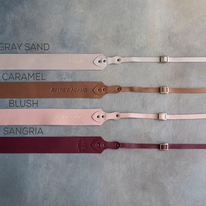 May include: Four leather camera straps in different colors: gray sand, caramel, blush, and sangria. Each strap has a metal buckle and is personalized with a name.