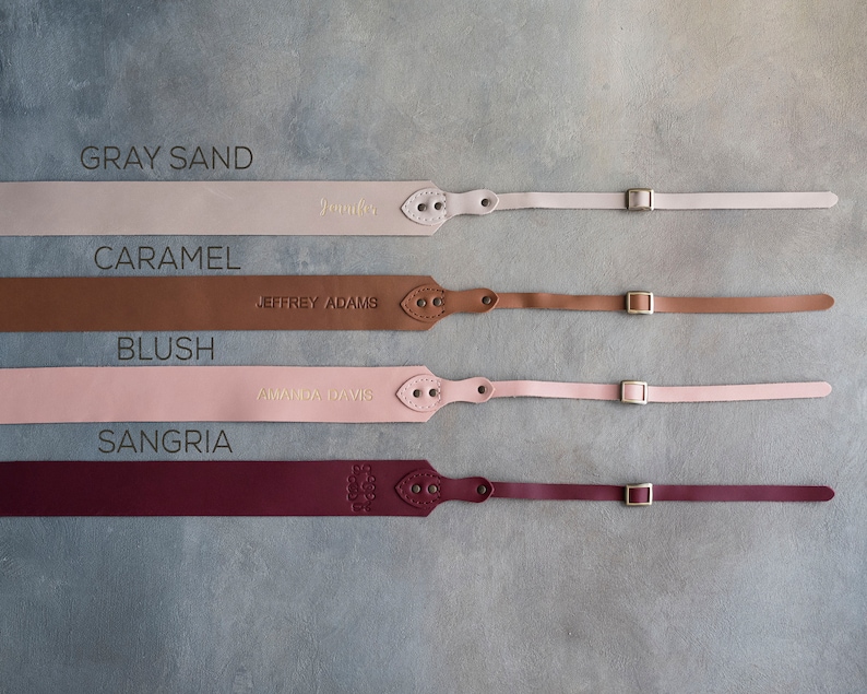 May include: Four leather camera straps in different colors: gray sand, caramel, blush, and sangria. Each strap has a metal buckle and is personalized with a name.