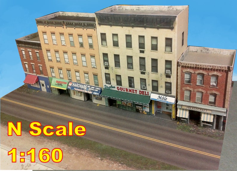 Peut inclure: Un ensemble de train miniature repr&eacute;sentant une rue de ville avec cinq b&acirc;timents, chacun avec une devanture de magasin. Les b&acirc;timents sont peints dans des tons de jaune, rouge et marron. La rue est pav&eacute;e d'asphalte et a des lignes jaunes au centre. Le texte "N Scale 1:160" est imprim&eacute; en bas de l'image.