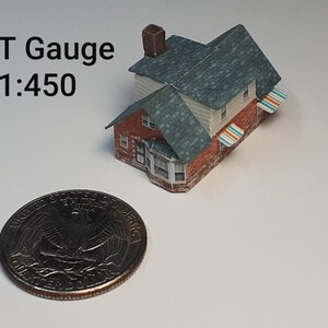May include: A small, red brick house with a green roof and white awnings. The house is about the size of a US quarter. The text "T Gauge 1:450" is visible in the image.