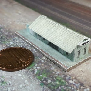 May include: A small model railway station, light green with a grey roof, labelled "ALLORA". The model sits on a rectangular base. A penny is next to the model for scale. The model is approximately 2.5 cm long.