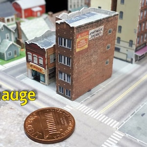 May include: A model railway layout featuring a T Gauge sign, a penny for scale, and several papercraft buildings, including a pizza shop and an apartment building with a sign that reads "Apartments For Rent".