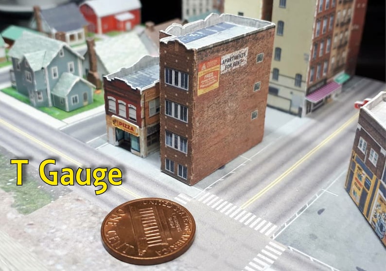 May include: A model railway layout featuring a T Gauge sign, a penny for scale, and several papercraft buildings, including a pizza shop and an apartment building with a sign that reads "Apartments For Rent".