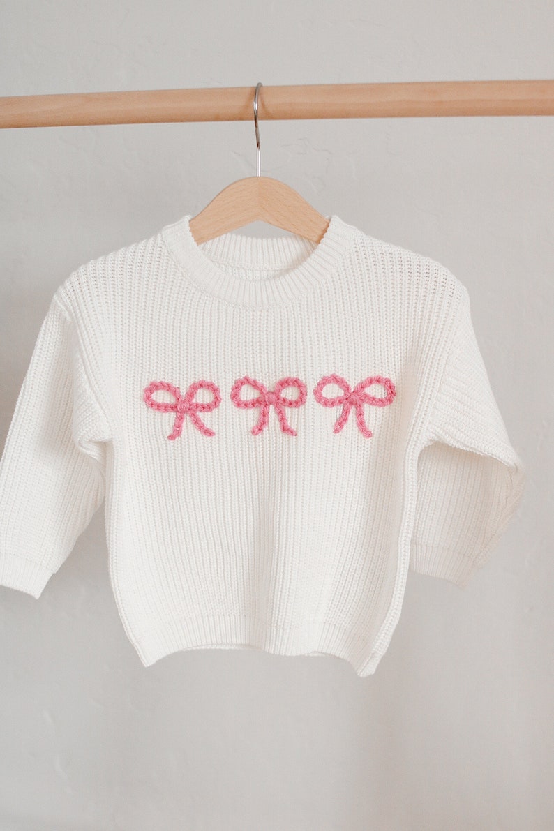 May include: White knitted sweater with a crew neck and pink embroidered bows on the front.