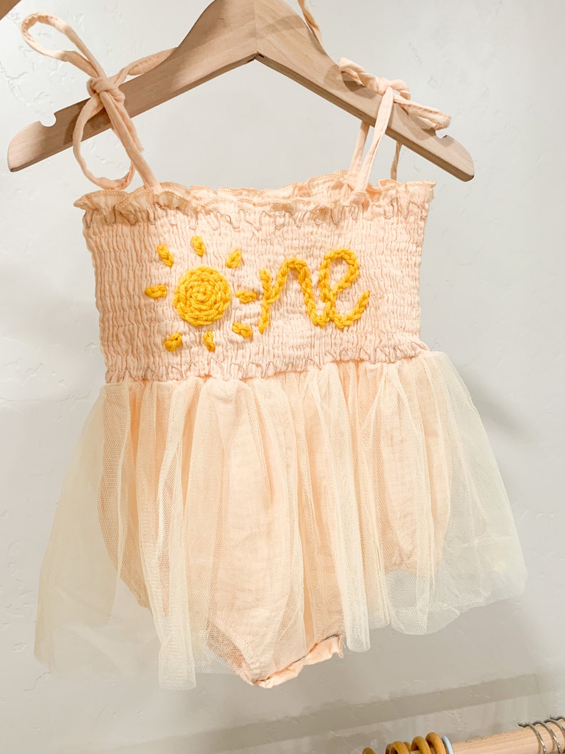 Op de afbeelding: Een perzikkleurige baby romper met een gele geborduurde zon en het woord "one". De romper heeft een tule rok.