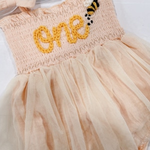 May include: A peach-colored tulle baby dress with a yellow knitted "one" and a bee applique. The dress has thin straps and a ruffled neckline.