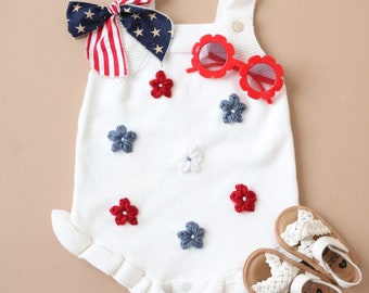 May include: White knitted baby romper with red, white, and blue flower embellishments. The romper has a ruffled hem and is paired with red sunglasses and white sandals.