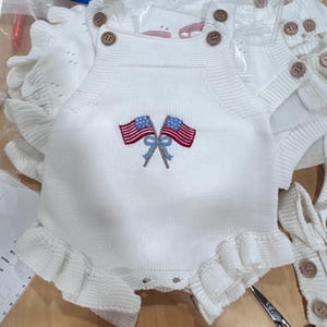 Puede incluir: Un pelele de punto blanco sin mangas con dos botones marrones en cada tirante. El centro del pecho muestra un gr&aacute;fico bordado de dos banderas estadounidenses cruzadas atadas con una cinta azul claro. Las aberturas de las piernas tienen volantes.