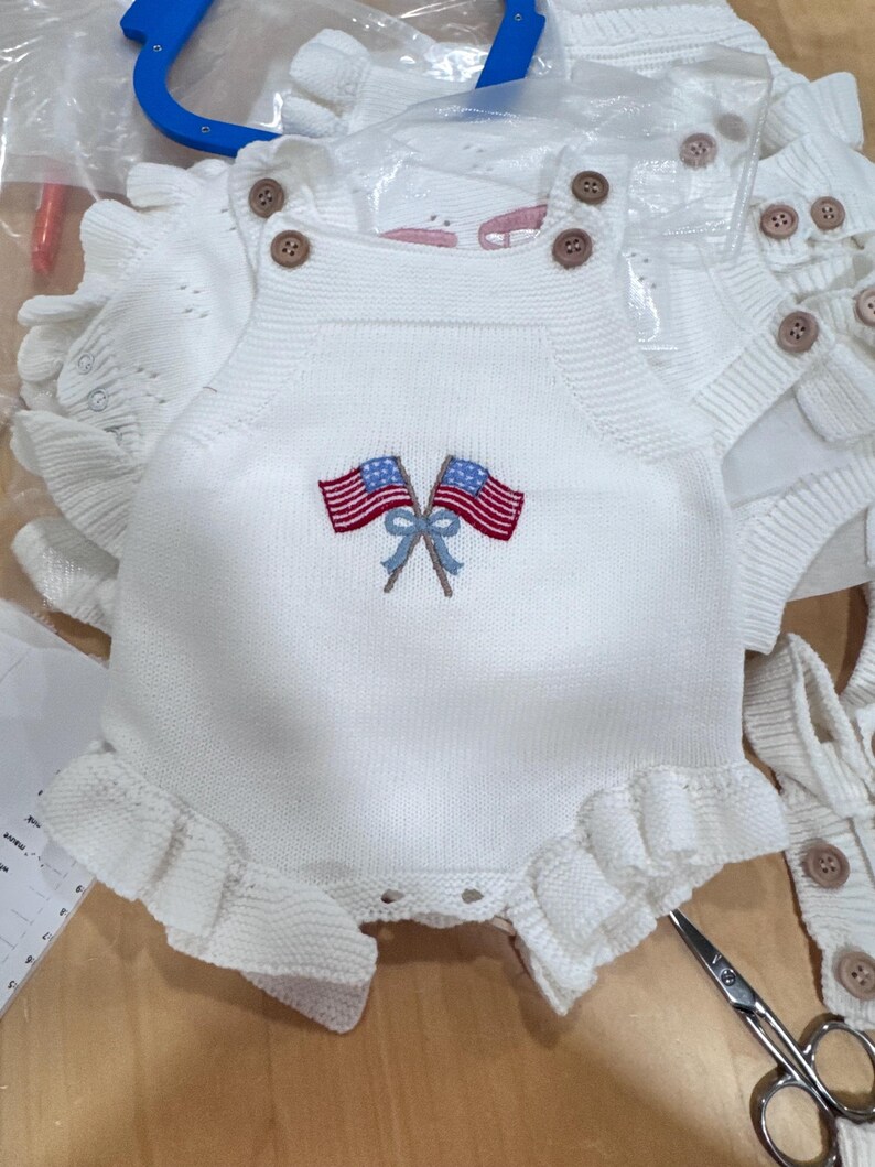 Puede incluir: Un pelele de punto blanco sin mangas con dos botones marrones en cada tirante. El centro del pecho muestra un gr&aacute;fico bordado de dos banderas estadounidenses cruzadas atadas con una cinta azul claro. Las aberturas de las piernas tienen volantes.