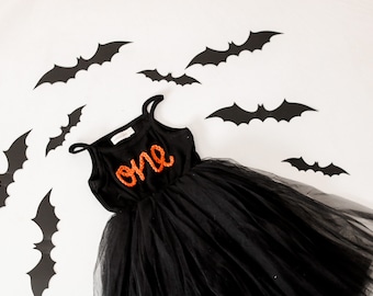 May include: A black tulle tutu dress with a black velvet bodice and the word "one" embroidered in orange thread. The dress is surrounded by black paper bats.