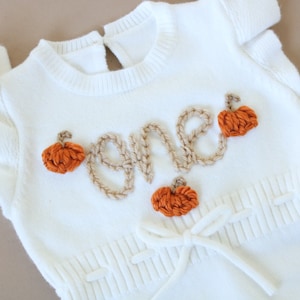 May include: White knitted baby onesie with the word "one" embroidered in brown yarn and decorated with orange felt pumpkins.