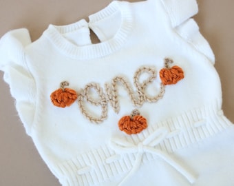May include: White knitted baby onesie with the word "one" embroidered in brown yarn and decorated with orange felt pumpkins.