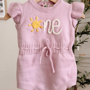 May include: A pink knitted baby onesie with a yellow sun and white embroidered "one" on the front. The onesie has a tie at the waist and wooden buttons at the bottom.