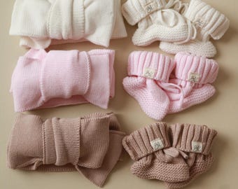Könnte beinhalten: Drei Paar gestrickte Babyschuhe in Weiß, Rosa und Braun. Jedes Paar hat eine Schleife oben und ein kleines Etikett mit dem Buchstaben "B" darauf.