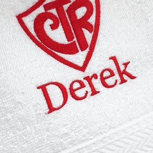 May include: A white towel featuring red embroidery. The design includes a shield-shaped emblem containing the letters CTR, positioned directly above the name Derek, which is stitched in a bold, red, sans-serif font on the textured fabric.