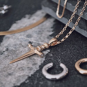 Pode incluir: Colar de corrente de ouro com um pingente de espada e uma pequena pedra transparente. O colar est&aacute; sobre uma superf&iacute;cie cinza com duas argolas de orelha douradas.