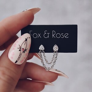 Peut inclure: Une paire de boucles d'oreilles en argent avec une pierre en forme de larme et une chaîne pendante. Les boucles d'oreilles sont présentées sur une carte noire avec le texte "Fox & Rose" imprimé en blanc.
