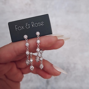 Puede incluir: Pendientes colgantes plateados con múltiples cristales transparentes. Los pendientes tienen un diseño delicado con un grupo de cristales en la parte superior y cristales en forma de lágrima en cascada. Se muestran con una tarjeta negra que dice "Fox & Rose".