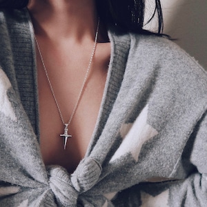 Pode incluir: Um cardig&atilde; cinza com padr&otilde;es de estrelas brancas amarrado na frente. O cardig&atilde; &eacute; usado sobre uma camiseta branca sem mangas e um colar prateado com um pingente de adaga.