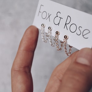Pode incluir: Três pares de brincos de corrente dourada com pequenas bolas nas pontas. Os brincos são exibidos em um cartão branco com o texto "Fox & Rose" impresso nele.