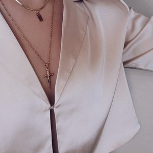 Pode incluir: Um close-up de uma pessoa vestindo uma camisa de cetim bege claro com um colar de ouro em camadas com um pingente de espada.