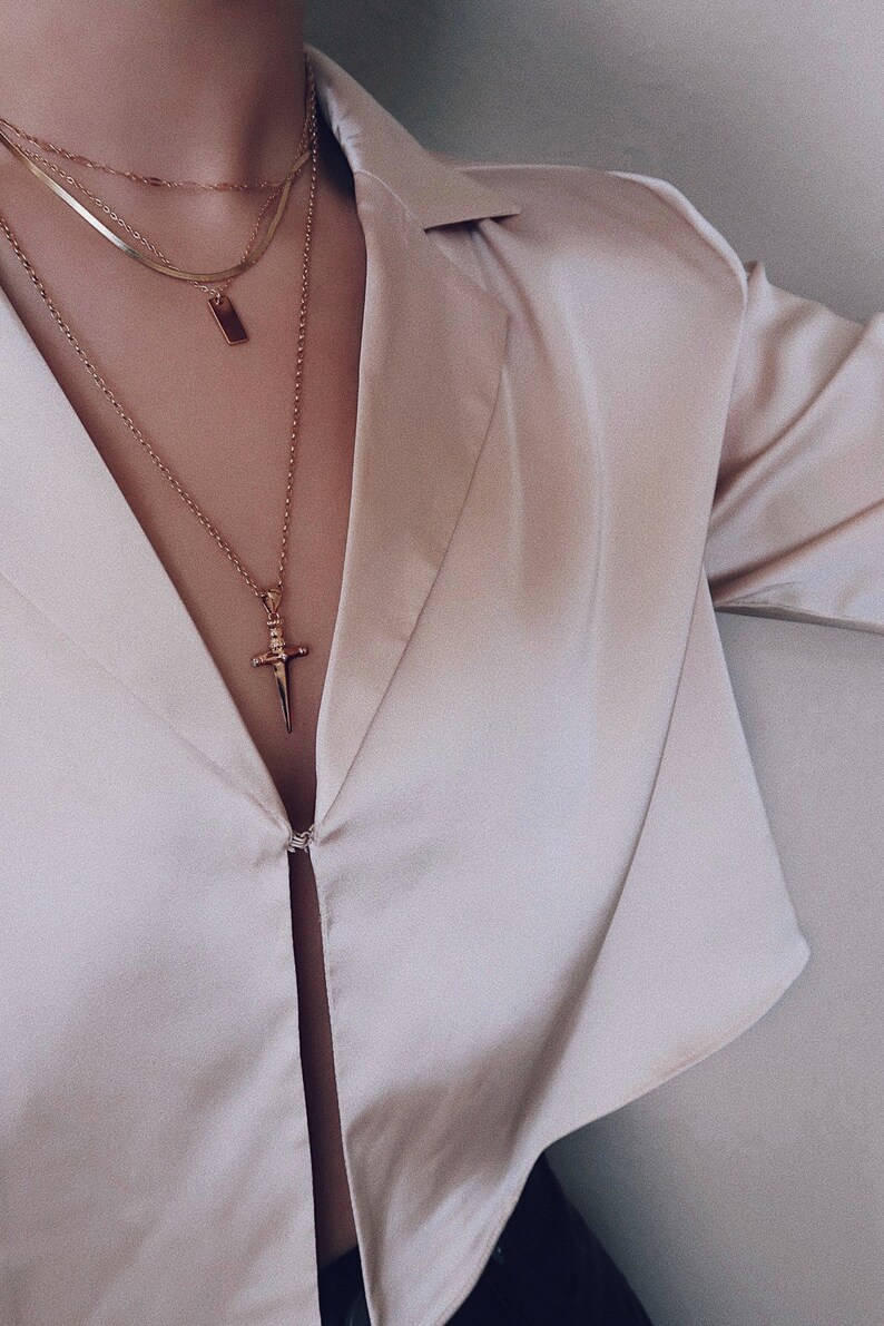 Pode incluir: Um close-up de uma pessoa vestindo uma camisa de cetim bege claro com um colar de ouro em camadas com um pingente de espada.
