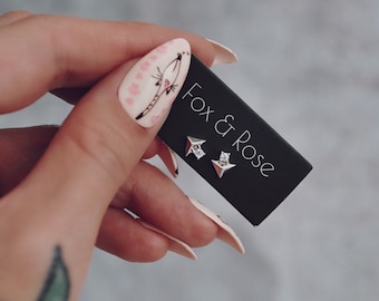 May include: A pair of silver star-shaped earrings with a small clear stone in the center. The earrings are on a black card with the text "Fox & Rose" printed in white.