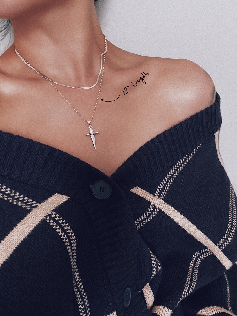Pode incluir: Um colar de corrente de prata com um pingente de espada. O colar tem 45,7 cm de comprimento e &eacute; usado por uma pessoa com um cardigan listrado preto e branco.