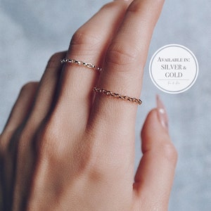 May include: Two gold chain rings are worn on a hand with a light skin tone. The rings are delicate, with a chain link design. The image also includes the text "AVAILABLE IN: SILVER & GOLD".