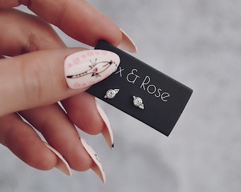 May include: A pair of small, silver-toned stud earrings with a clear, round gemstone center, displayed on a black card. The card reads "Fox & Rose" in white text. The image is held by a hand with long, manicured nails.