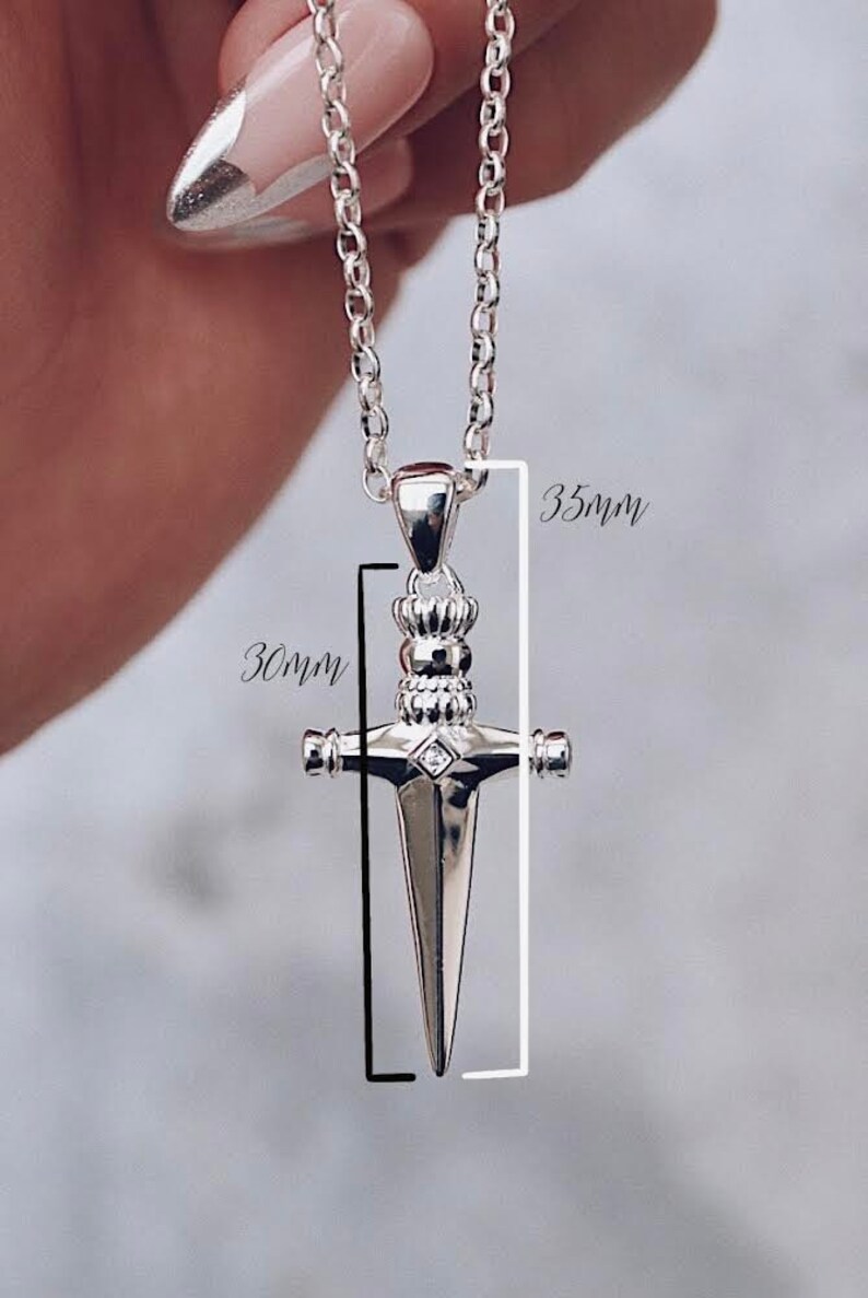 Pode incluir: Colar com pingente de prata com um design de adaga detalhado. O pingente mede 30 mm de largura e 35 mm de comprimento.