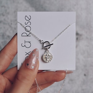 May include: A silver necklace with a toggle clasp and a round coin pendant. The coin has a textured design and a raised image of a woman's face. The necklace is displayed on a white card with the text "Fox & Rose" printed on it.
