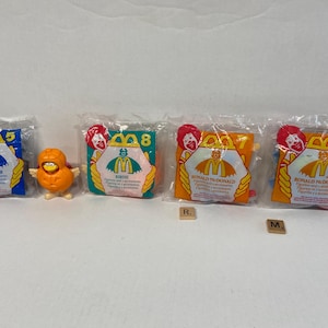 May include: A collection of McDonald's Happy Meal toys, including a small orange bird figure and several sealed bags. The bags are blue, green, and orange, featuring Ronald McDonald and the McDonald's logo.
