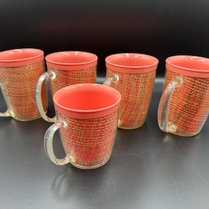 May include: A set of five clear glass mugs featuring a coral-colored rim and handle, with a textured, woven-style insert in shades of orange and red visible through the transparent walls of each individual drinking vessel.