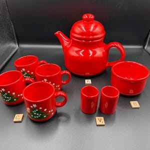May include: A bright red ceramic tea set consisting of a teapot with a lid, three mugs decorated with green Christmas tree graphics and white dots, two small cylindrical containers, and one bowl, accompanied by small wooden letter tiles.