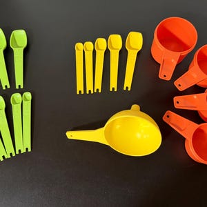May include: A collection of colorful measuring tools, including green measuring spoons, yellow measuring spoons, yellow measuring cups, and orange measuring cups. The tools are arranged on a dark surface, ideal for baking and cooking.