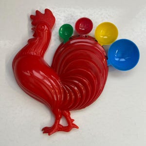 May include: Red rooster-shaped measuring spoons with four colorful measuring cups attached to the tail. The cups are red, green, yellow, and blue.