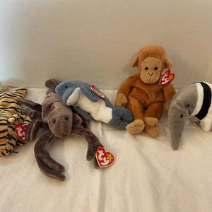 May include: A collection of plush animal toys, including a tiger with black stripes, a brown spider, a blue and white dolphin, a brown monkey, and a gray anteater with a black stripe. Each toy has a red tag.