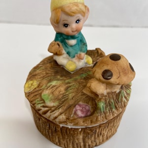 May include: A ceramic trinket box featuring a seated gnome figurine with a yellow hat and teal shirt. The box is brown with a mushroom and grass design on the lid. The box is approximately 3 inches in diameter.