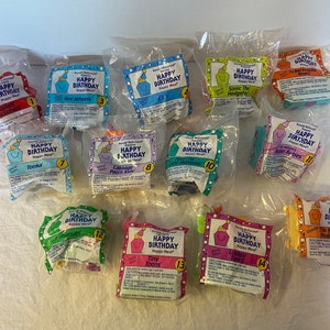 May include: A collection of sealed McDonald's Happy Meal toys, each in a clear plastic bag. The bags feature the text "Happy Birthday" and various cartoon characters, including Ronald McDonald, E.T., and Looney Tunes characters. The toys are numbered.