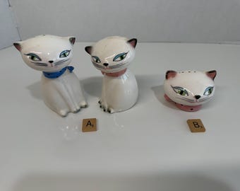 May include: Three white ceramic cat figurines with black accents, green eyes, and pink noses. One cat wears a blue scarf, another a pink collar, and the third has a pink base. These decorative cat figurines are likely salt and pepper shakers.