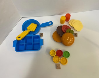 Puede incluir: Una bandeja de plástico azul con nueve compartimentos, una espátula amarilla, una sartén azul y un juego de alimentos de plástico de colores. Los alimentos incluyen una hamburguesa, un pan y una variedad de ingredientes. También hay un juego de números de plástico de colores.