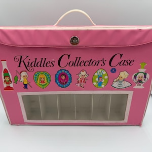 May include: A pink rectangular Kiddles Collector's Case with a white handle and a metal latch. The front features the text Kiddles Collector's Case above colorful illustrations of various doll characters and a clear rectangular viewing window.