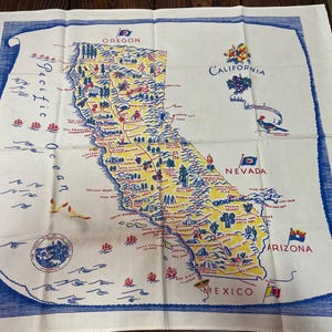 May include: White cotton tea towel featuring a vintage map of California, with surrounding states and the Pacific Ocean. The map is in yellow, blue, and red, with state names and illustrations.