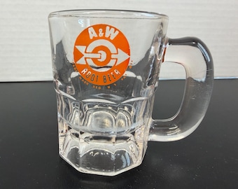 May include: Clear glass A&W Root Beer mug with a handle. The mug features an orange and white logo with the text "A&W Root Beer" in orange. The mug has a faceted design on the bottom half.