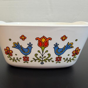 May include: White rectangular bowl with handles, decorated with blue birds, orange, yellow, and red flowers. The bowl has a vintage, folk art style design. The bowl is likely made of ceramic or a similar material.