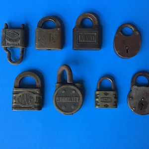 May include: Collection of eight antique padlocks in various shapes and sizes, arranged on a blue background. The padlocks are made of metal and show signs of age and wear. Some have text, including "TOWNLEY" and "VICTORY".