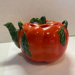 May include: A ceramic teapot shaped like a large tomato, with a glossy orange finish. Green leaves and a stem form the handle and lid. The spout and additional handle are also green.